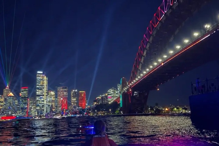 Person kayaking at night near lit-up city skyline and bridge.