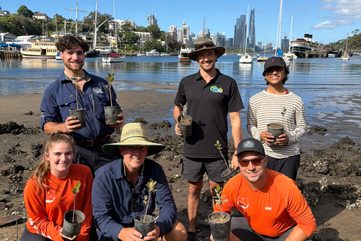 Group of seven people holding plants by a waterfront, wearing outdoor gear.