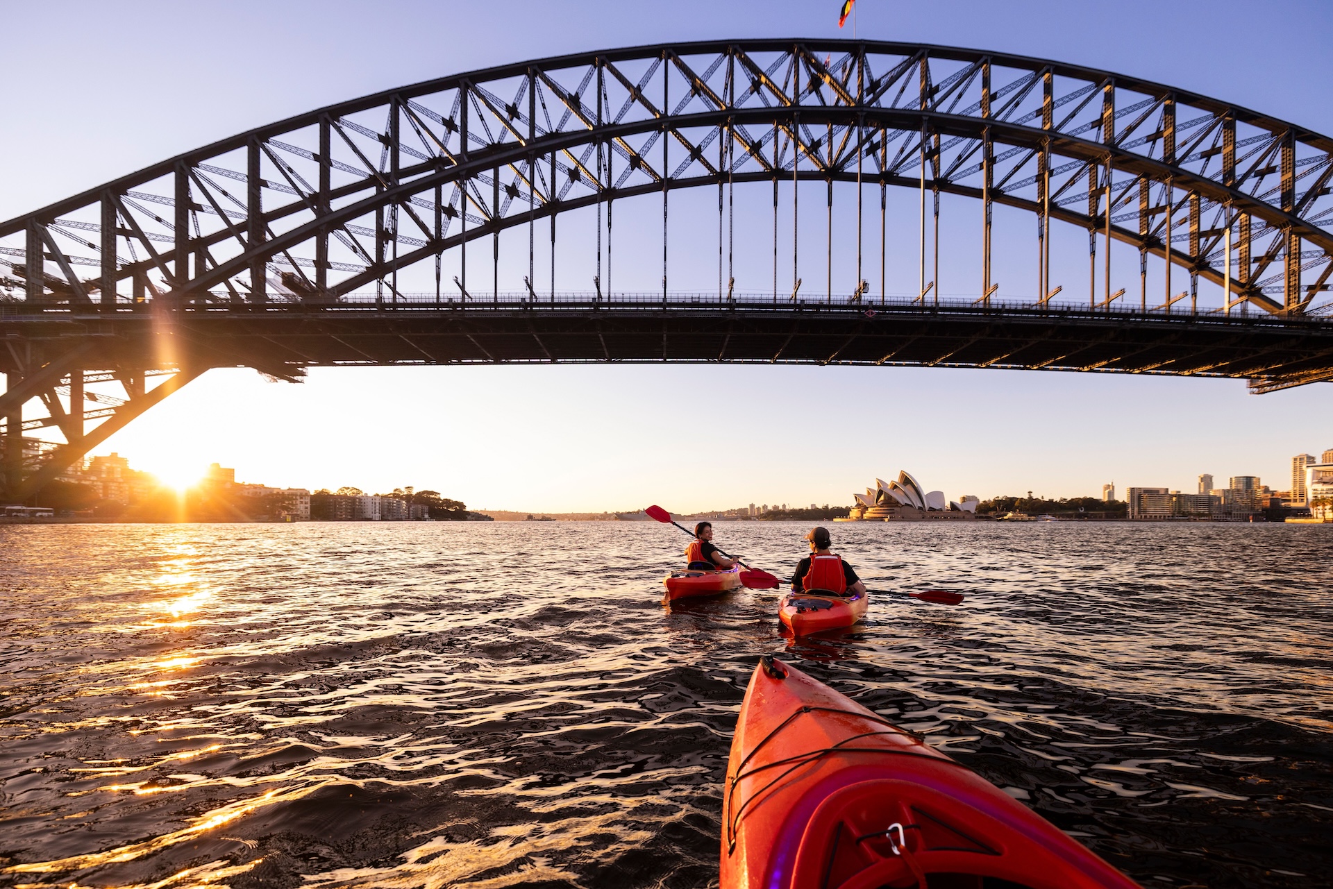 Two people kayaking under a large bridge with a cityscape at sunset.
