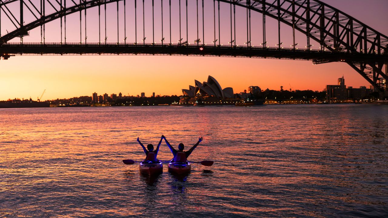 Two people kayaking under a bridge at sunset with city skyline in the background.