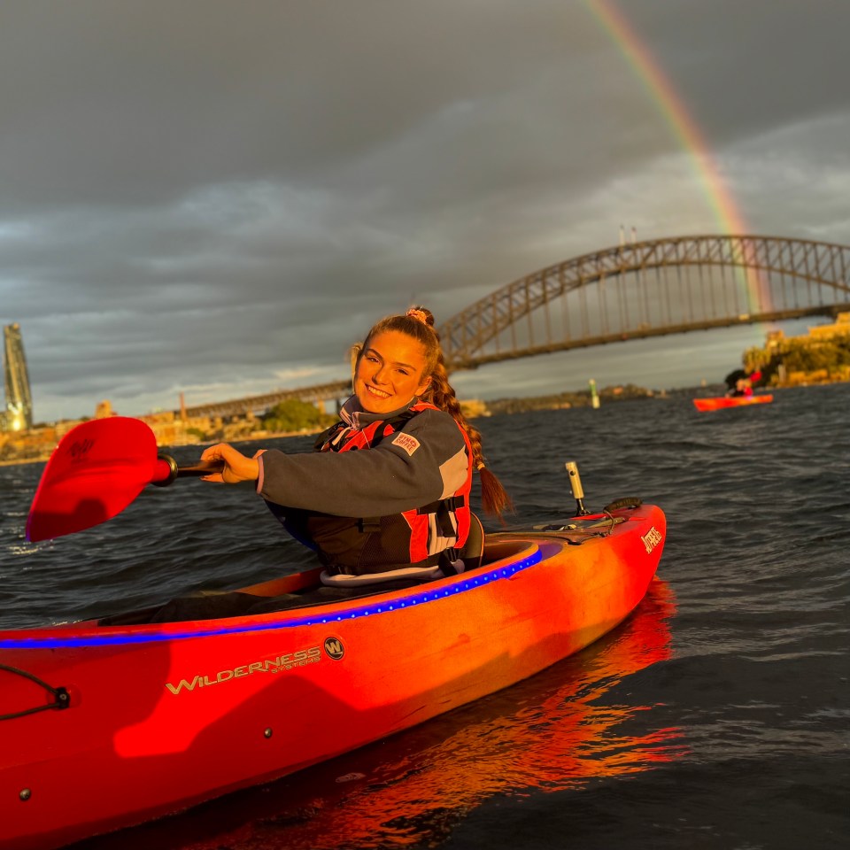Person in red kayak on water, with a rainbow and bridge in the background.