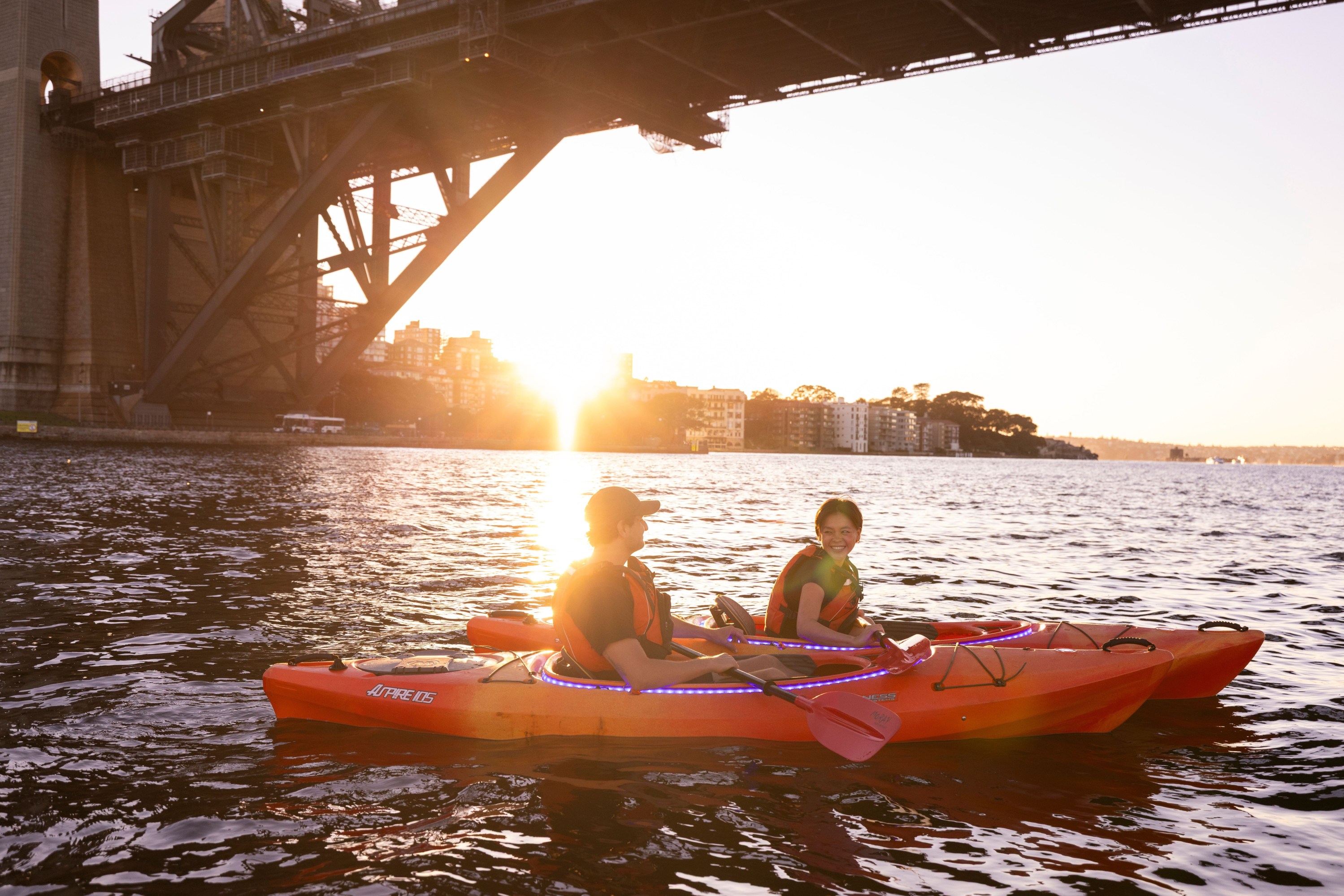 Two people kayaking under a bridge at sunset.
