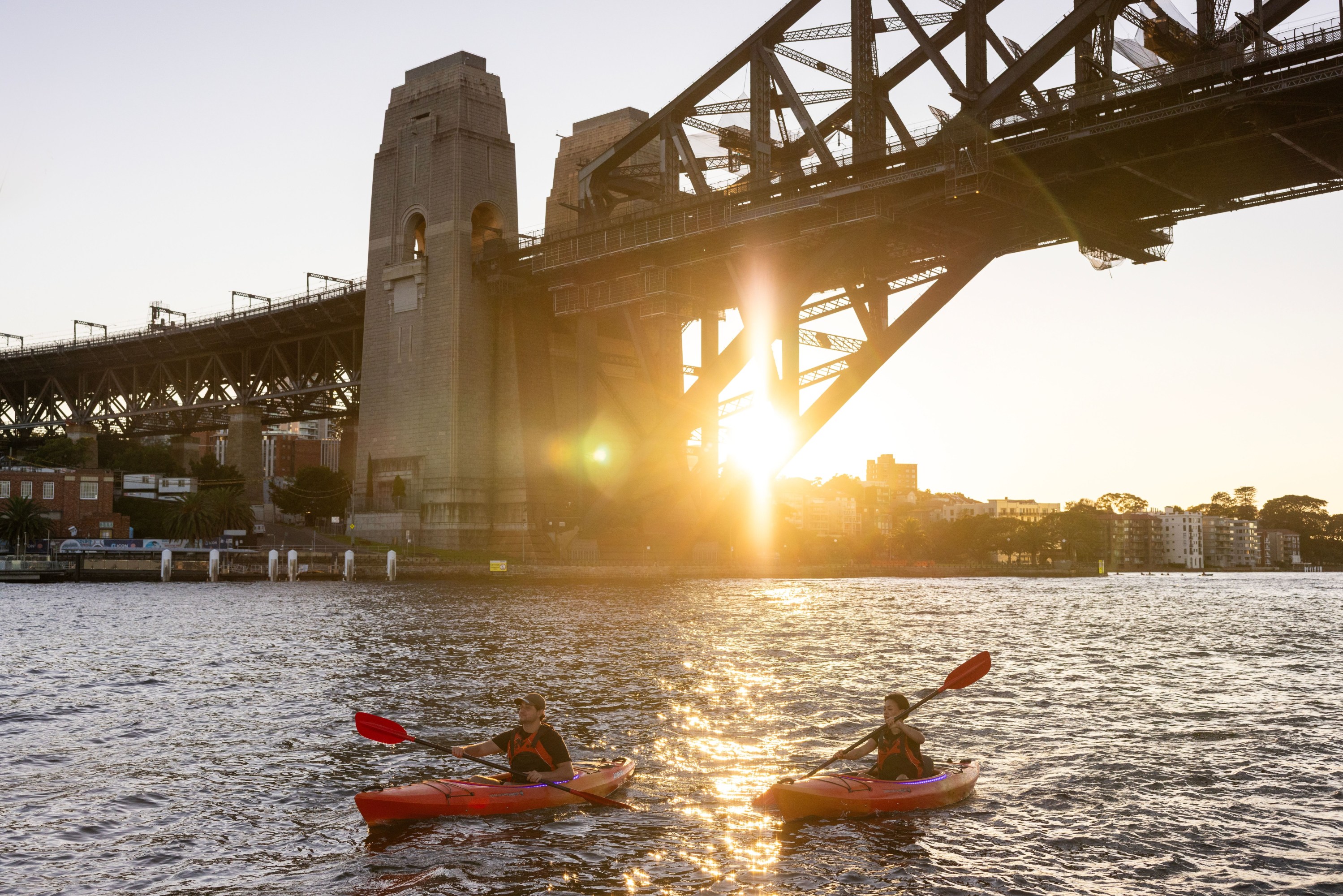 Two kayakers on water near a bridge at sunset.